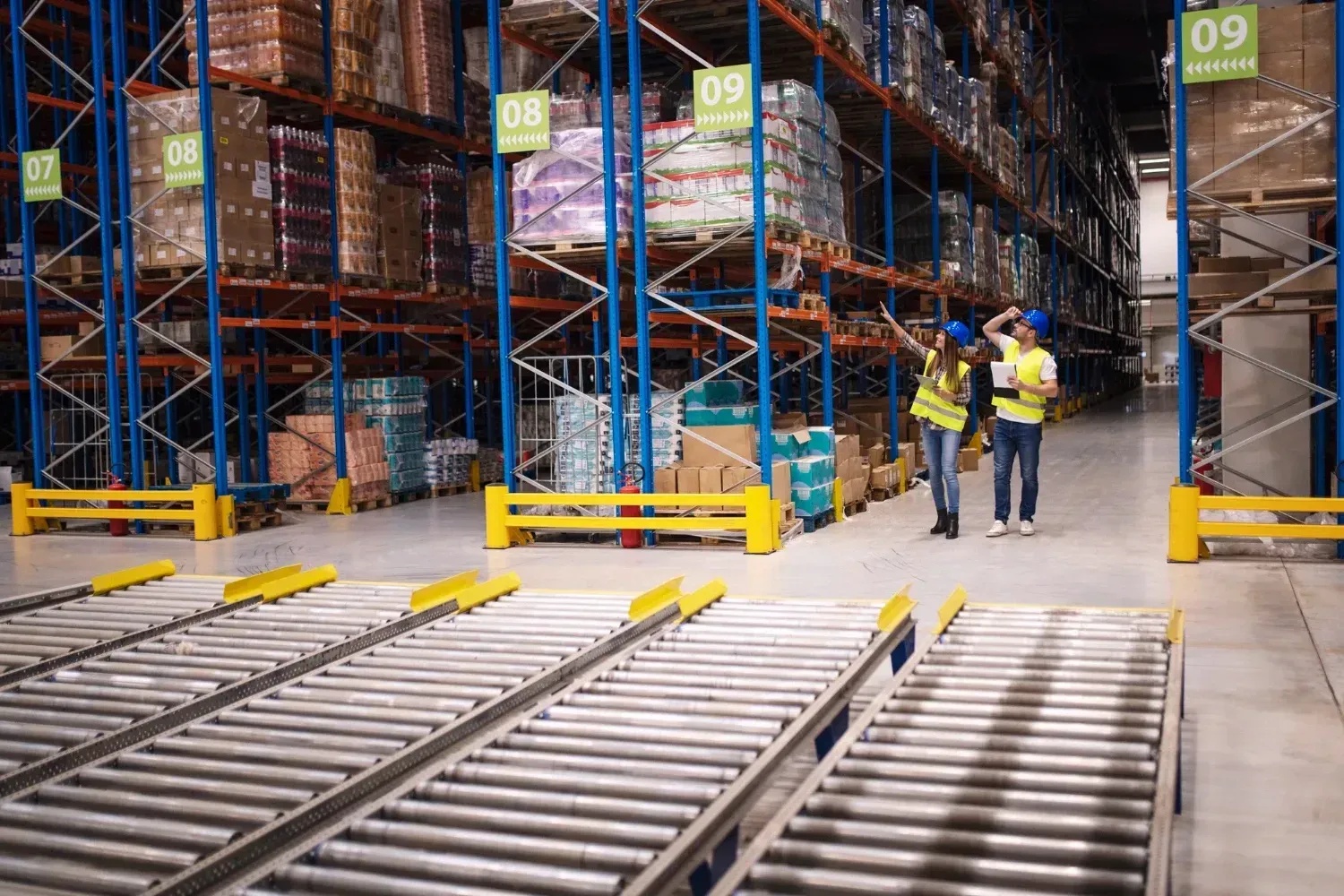 Two workers in hi-vis vests checking pallet racking above roller conveyors